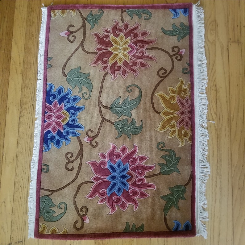 May include: A beige rug with a floral pattern featuring pink, blue, and yellow flowers. The rug has a white fringe border and a red border around the edge.