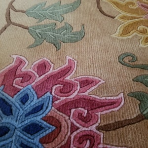 May include: Close-up of a beige rug with a floral design. The rug features pink, blue, and green flowers with intricate details. The background is a light beige color.
