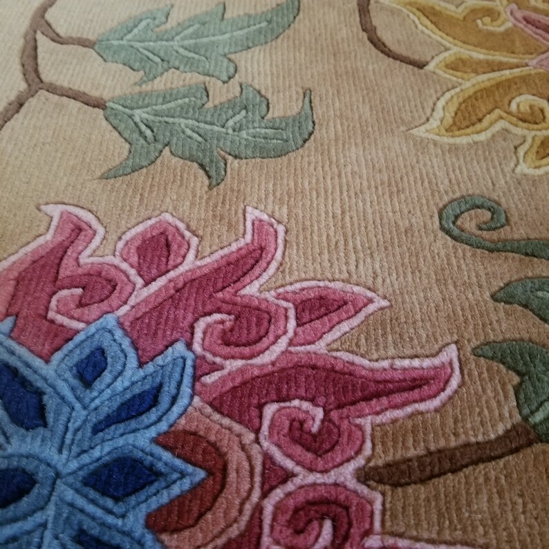 May include: Close-up of a beige rug with a floral design. The rug features pink, blue, and green flowers with intricate details. The background is a light beige color.