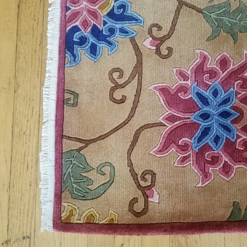 May include: A beige rug with a floral pattern featuring pink, blue, and green flowers. The rug has a fringe border and a solid pink edge.