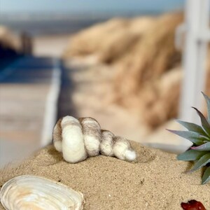 Könnte beinhalten: Eine Filz-Muscheln-Skulptur in Braun- und Cremetönen liegt auf Sand, neben einer weißen Muschel. Im Hintergrund eine verschwommene Strandszene. Der Künstlername FilzatelierEla ist unten links zu sehen.