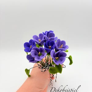 K&ouml;nnte beinhalten: Ein Strau&szlig; handgefertigter violetter Blumen mit gr&uuml;nen Bl&auml;ttern und gelben Zentren. Die Blumen sind mit einem schwarz-wei&szlig; gestreiften Band gebunden. Der Text "Dekobeispiel mit 3 Veilchenstr&auml;u&szlig;en" ist sichtbar.