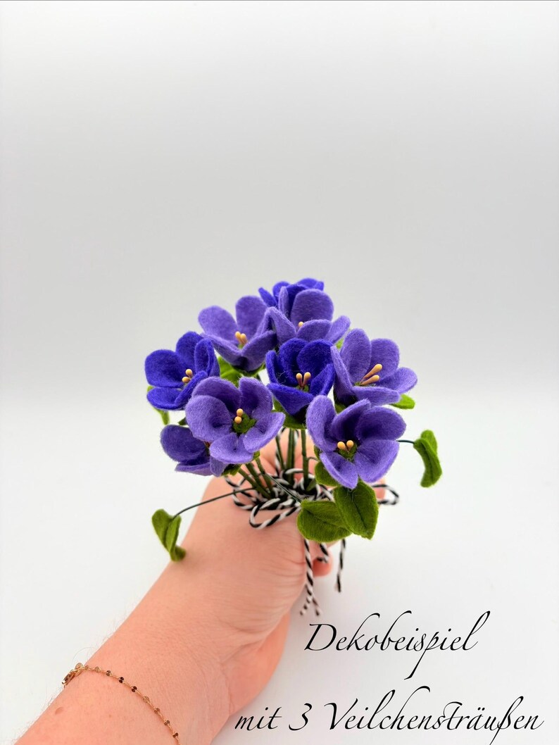 K&ouml;nnte beinhalten: Ein Strau&szlig; handgefertigter violetter Blumen mit gr&uuml;nen Bl&auml;ttern und gelben Zentren. Die Blumen sind mit einem schwarz-wei&szlig; gestreiften Band gebunden. Der Text "Dekobeispiel mit 3 Veilchenstr&auml;u&szlig;en" ist sichtbar.