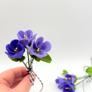 K&ouml;nnte beinhalten: Ein kleines Bouquet handgefertigter Veilchen aus Filz in Lila- und Gr&uuml;nt&ouml;nen. Die Blumen haben gelbe Zentren und sind mit einer schwarz-wei&szlig;en Schnur zusammengebunden. Der Hintergrund ist schlicht wei&szlig;.