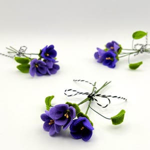 K&ouml;nnte beinhalten: Drei kleine Str&auml;u&szlig;e k&uuml;nstlicher Veilchen. Jeder Strau&szlig; zeigt leuchtend violette Bl&uuml;ten mit gelben Zentren und gr&uuml;nen Bl&auml;ttern. Die Stiele sind mit schwarz-wei&szlig;em Garn gebunden, was eine zarte und charmante &Auml;sthetik erzeugt.