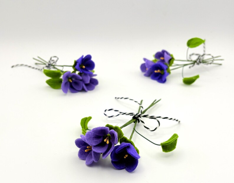 K&ouml;nnte beinhalten: Drei kleine Str&auml;u&szlig;e k&uuml;nstlicher Veilchen. Jeder Strau&szlig; zeigt leuchtend violette Bl&uuml;ten mit gelben Zentren und gr&uuml;nen Bl&auml;ttern. Die Stiele sind mit schwarz-wei&szlig;em Garn gebunden, was eine zarte und charmante &Auml;sthetik erzeugt.