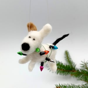Könnte beinhalten: Handgefertigter weißer Filzhunde-Ornament mit schwarzer Nase und Schwanz sowie braunen Ohren. Der Hund ist mit einer Lichterkette in bunten Farben geschmückt. Eine festliche Weihnachtsdekoration.