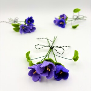 K&ouml;nnte beinhalten: Nahaufnahme von mehreren Filz-Veilchen-Blumenstr&auml;u&szlig;en. Die Blumen sind tiefviolett mit gelben Zentren und gr&uuml;nen Bl&auml;ttern. Jeder Strau&szlig; ist mit einer schwarz-wei&szlig;en Schnur gebunden. Der Hintergrund ist schlicht wei&szlig;.