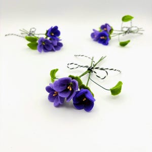 K&ouml;nnte beinhalten: Drei kleine Str&auml;u&szlig;e aus Filzblumen mit leuchtend violetten Bl&uuml;tenbl&auml;ttern und gr&uuml;nen Bl&auml;ttern. Jeder Strau&szlig; ist mit schwarz-wei&szlig;em Garn gebunden. Der Text "Filzatelier Ela" befindet sich in der unteren linken Ecke.