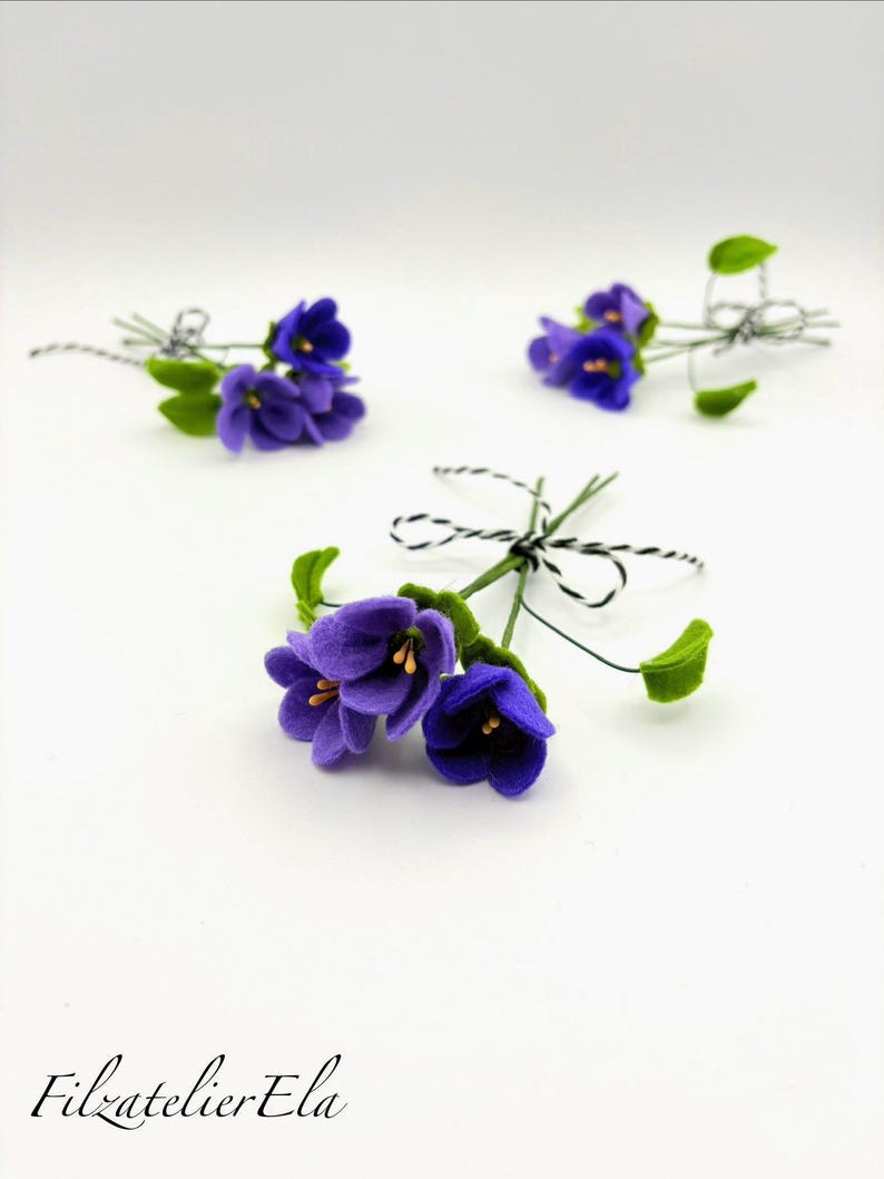 K&ouml;nnte beinhalten: Drei kleine Str&auml;u&szlig;e aus Filzblumen mit leuchtend violetten Bl&uuml;tenbl&auml;ttern und gr&uuml;nen Bl&auml;ttern. Jeder Strau&szlig; ist mit schwarz-wei&szlig;em Garn gebunden. Der Text "Filzatelier Ela" befindet sich in der unteren linken Ecke.