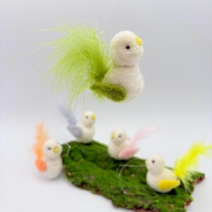 Könnte beinhalten: Handgefertigte Filz-Vogelornamente mit bunten Federschwänzen. Die Vögel sind weiß mit gelben Schnäbeln und Akzenten, die auf einem Stück grünen Moos sitzen. Das Bild enthält den Text "Filzatelier Ela".