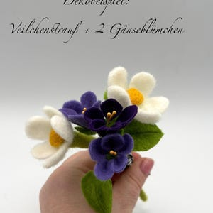 K&ouml;nnte beinhalten: Ein handgefertigter Filzblumenstrau&szlig; mit wei&szlig;en und violetten Bl&uuml;ten mit gelben Zentren und gr&uuml;nen Bl&auml;ttern. Der Strau&szlig; wird in einer Hand gehalten. Der Text im Bild lautet "Dekobeispiel: Veilchenstrau&szlig; + 2 G&auml;nsebl&uuml;mchen".