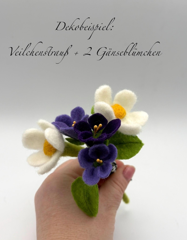 K&ouml;nnte beinhalten: Ein handgefertigter Filzblumenstrau&szlig; mit wei&szlig;en und violetten Bl&uuml;ten mit gelben Zentren und gr&uuml;nen Bl&auml;ttern. Der Strau&szlig; wird in einer Hand gehalten. Der Text im Bild lautet "Dekobeispiel: Veilchenstrau&szlig; + 2 G&auml;nsebl&uuml;mchen".