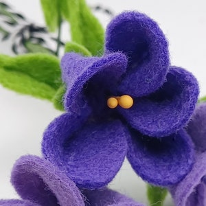 K&ouml;nnte beinhalten: Nahaufnahme von handgefertigten Veilchenbl&uuml;ten aus Filz mit lila Bl&uuml;tenbl&auml;ttern, gelben Staubgef&auml;&szlig;en und gr&uuml;nen Bl&auml;ttern. Die Blumen sind detailliert und wirken weich, mit einer strukturierten Oberfl&auml;che. Der Hintergrund ist wei&szlig;.