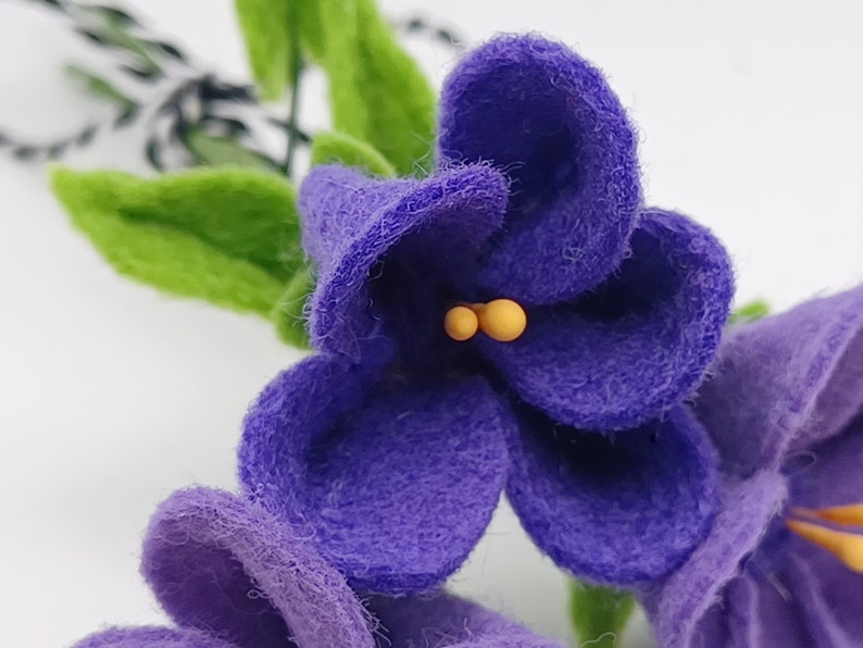 K&ouml;nnte beinhalten: Nahaufnahme von handgefertigten Veilchenbl&uuml;ten aus Filz mit lila Bl&uuml;tenbl&auml;ttern, gelben Staubgef&auml;&szlig;en und gr&uuml;nen Bl&auml;ttern. Die Blumen sind detailliert und wirken weich, mit einer strukturierten Oberfl&auml;che. Der Hintergrund ist wei&szlig;.