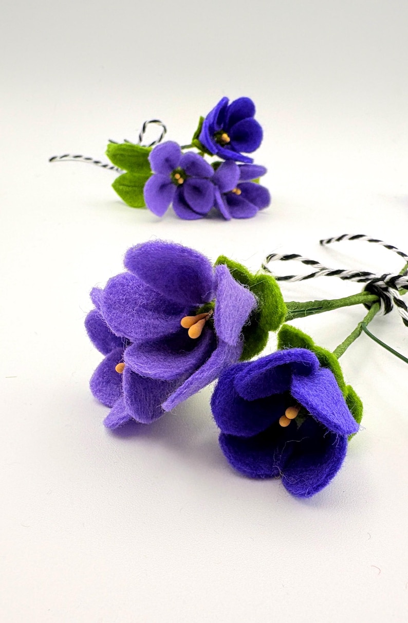 K&ouml;nnte beinhalten: Zwei handgefertigte Veilchenblumenstr&auml;u&szlig;e aus Filz. Die Bl&uuml;ten sind tiefviolett mit gelben Zentren und gr&uuml;nen Bl&auml;ttern. Die Stiele sind gr&uuml;n mit einer schwarz-wei&szlig;en Schnur. Der Hintergrund ist wei&szlig;.