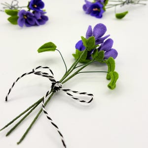 K&ouml;nnte beinhalten: Handgefertigte Veilchenbl&uuml;ten aus Filz mit gr&uuml;nen Bl&auml;ttern und Stielen, gebunden mit schwarz-wei&szlig;em Garn. Die lila Bl&uuml;ten sind in kleinen Str&auml;u&szlig;en angeordnet.