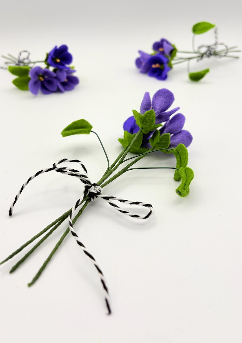 K&ouml;nnte beinhalten: Handgefertigte Veilchenbl&uuml;ten aus Filz mit gr&uuml;nen Bl&auml;ttern und Stielen, gebunden mit schwarz-wei&szlig;em Garn. Die lila Bl&uuml;ten sind in kleinen Str&auml;u&szlig;en angeordnet.