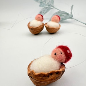 Könnte beinhalten: Handgefertigte Babyfiguren aus Filz in Walnussschalen. Jedes Baby hat eine rote Mütze und ein pfirsichfarbenes Gesicht, das auf weißer Wolle ruht. Der Text "Filzatelier Ela" ist enthalten.