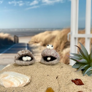 Könnte beinhalten: Zwei Filzmuschelfiguren mit weißen Augen und einer Perle. Die Figuren befinden sich auf einem Sandbett mit einer Muschel und kleinen Steinen. Der Hintergrund zeigt eine Strandszene mit blauem Himmel und Meer.