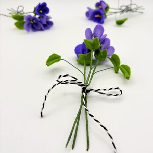 K&ouml;nnte beinhalten: Mehrere kleine Str&auml;u&szlig;e k&uuml;nstlicher Veilchen. Jeder Strau&szlig; zeigt leuchtend violette Bl&uuml;ten mit gr&uuml;nen Bl&auml;ttern und Stielen. Die Stiele sind mit schwarz-wei&szlig;em Bindfaden zusammengebunden. Der Hintergrund ist schlicht wei&szlig;.