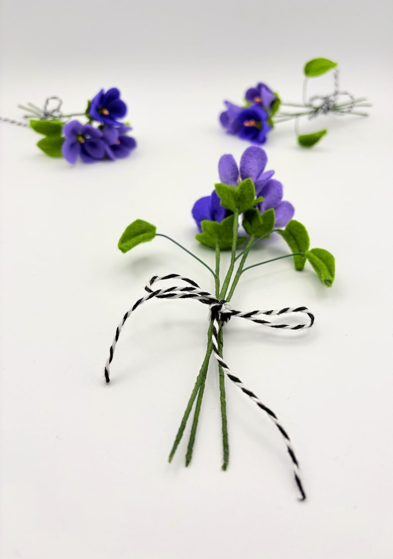 K&ouml;nnte beinhalten: Mehrere kleine Str&auml;u&szlig;e k&uuml;nstlicher Veilchen. Jeder Strau&szlig; zeigt leuchtend violette Bl&uuml;ten mit gr&uuml;nen Bl&auml;ttern und Stielen. Die Stiele sind mit schwarz-wei&szlig;em Bindfaden zusammengebunden. Der Hintergrund ist schlicht wei&szlig;.