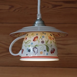 Könnte beinhalten: Eine weiße Teetasse aus Keramik mit Blumenmuster wird als Hängelampe verwendet. Die Teetasse hat ein rotes, gelbes und blaues Blumenmuster. Die Leuchte ist an einem weißen Kabel aufgehängt und hat eine weiße Untertasse.