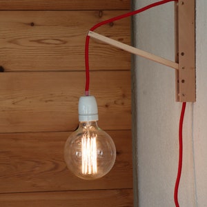 Könnte beinhalten: Ein rotes Kabel hängt an einer Wandhalterung aus Holz und verbindet sich mit einer klaren Glühbirne aus Glas mit einer weißen Keramikfassung. Die Glühbirne ist eingeschaltet und beleuchtet den Raum.