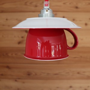 Könnte beinhalten: Eine rote Keramiktasse mit einem weißen Unterteller, umgewandelt in eine Hängelampe. Die Tasse hat einen Henkel und einen weißen Rand. Die Leuchte hängt an einer weißen Decke.