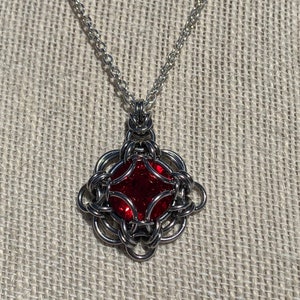 May include: A silver chain necklace with a red gemstone pendant. The pendant is a square shape with a woven design.