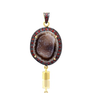 Puede incluir: Un colgante de plata con una piedra de ágata marrón engastada en un marco de metal negro con piedras preciosas rojas alrededor del borde. El colgante tiene una cadena de oro unida a la parte inferior.