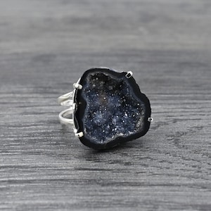 May include: A silver ring with a dark blue geode stone and a sparkling interior. The geode has a black border and is set on a double-banded silver ring. The ring is displayed on a grey wooden surface.
