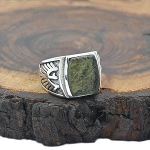 Puede incluir: Un anillo de plata con una piedra cuadrada de color verde oliva. El anillo tiene un diseño detallado en los laterales, con un motivo estilizado de pájaro o águila. El anillo está sobre una superficie de madera oscura.