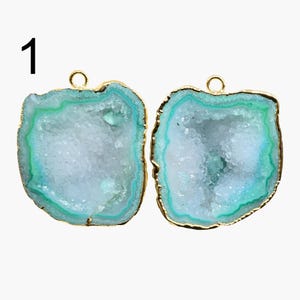 Puede incluir: Dos colgantes de amatista azul claro con bucles de metal dorado. La amatista tiene un borde verde y un interior blanco con una textura cristalina.