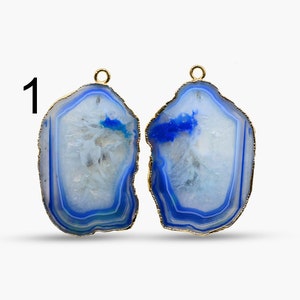 Puede incluir: Dos rebanadas de ágata azul y blanca con ribete dorado y un lazo dorado en la parte superior. Las rebanadas de ágata tienen una forma irregular y un aspecto natural y orgánico.