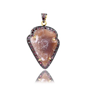 Puede incluir: Un colgante de plata con una piedra de ágata marrón engastada en un marco de metal negro. La piedra está rodeada por un borde de pequeñas piedras preciosas rojas.