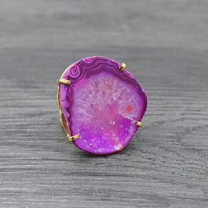 May include: A gold-coloured ring with a polished, oval agate stone. The agate has concentric bands of deep purple, magenta, and light pink. The stone is approximately 3.8 cm in diameter.