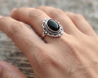 May include: A silver ring featuring an oval-shaped black stone set in a decorative metal frame with small circular accents along the edge. The band has a textured, beaded appearance, and the setting is centered on the finger.