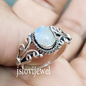 May include: A silver ring with a white moonstone set in a delicate, ornate design. The ring is shown on a hand.
