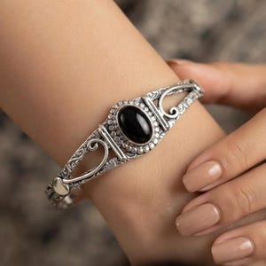 Può includere: Un bracciale rigido in argento con una pietra nera ovale incastonata al centro. La fascia metallica presenta intricati motivi a rilievo e un bordo perlato attorno alla pietra, con dettagli traforati che si estendono lungo i lati del bracciale.
