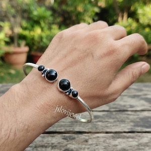 May include: A silver cuff bracelet featuring two open ends, each adorned with a cluster of smooth, round black stones. The metal band has a polished finish, and the text jslovijewel is visible on the surface of the silver near the stones.