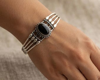 May include: A silver cuff bracelet featuring a central oval black stone set in a decorative frame. The band consists of four parallel silver wires that converge at the sides, accented with small silver bead details and twisted wire elements.