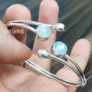 Puede incluir: Un brazalete de plata con dos piedras de luna. El brazalete está abierto y tiene un diseño sencillo.