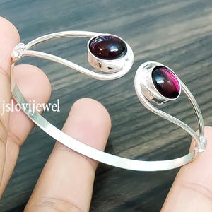 May include: A silver-toned open cuff bracelet featuring two oval, dark red gemstones. The gemstones are set in a bezel setting, and the bracelet has a smooth, polished finish. The bracelet is held by a hand.