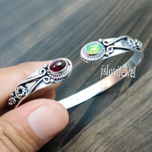 May include: Silver bangle bracelet with a red and green gemstone accent. The bracelet is open and has a delicate design. The text "jslovijewel" is visible on the bracelet.
