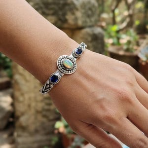 May include: A silver cuff bracelet featuring a central oval-shaped iridescent opal cabochon surrounded by a beaded border, flanked by two smaller oval-shaped deep blue faceted gemstones, all set within a decorative metal frame with intricate detailing.