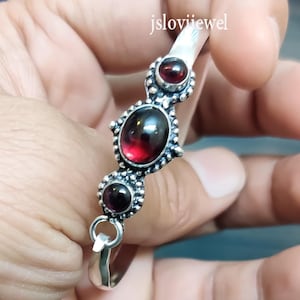 Pode incluir: Uma pulseira de prata com três pedras preciosas vermelhas escuras, em forma oval. As pedras estão engastadas em engastes de prata oxidada. A pulseira é segurada em uma mão, com o texto "jsloviijewel" visível.