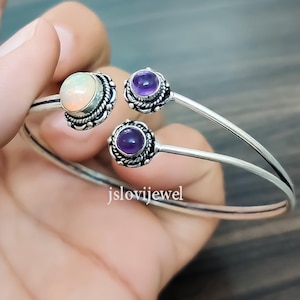 Può includere: Due braccialetti d'argento con dettagli decorativi. Un braccialetto presenta una pietra preziosa di opale bianco, mentre l'altro presenta due pietre preziose di ametista viola.