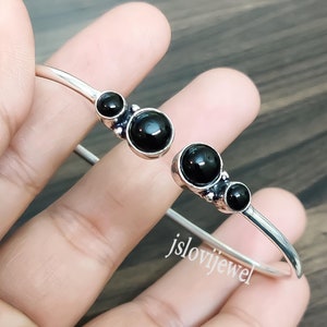 May include: A silver-toned cuff bracelet featuring four polished black onyx stones. The bracelet has an open design with the stones clustered in pairs at each end. The jewelry is displayed against a wooden surface.