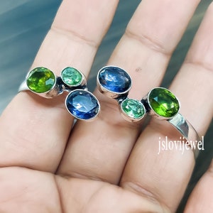 May include: A silver ring with a unique design featuring multiple green and blue gemstones. The ring is shown on a hand.